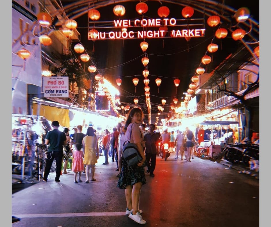 Cổng chợ đêm treo nhiều đèn lồng đỏ, bảng đèn chữ Welcome to Phu Quoc Night Market, một cô gái đeo ba lô đứng giữa đường quay lại nhìn máy ảnh, khách đi dạo giữa hai dãy hàng sáng đèn