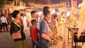 Hai người lớn tuổi và hai bé trai đứng xem một quầy nướng trên con phố lát gạch, phía sau treo cờ và dây đèn, cờ đề chữ Chợ Vui Phết