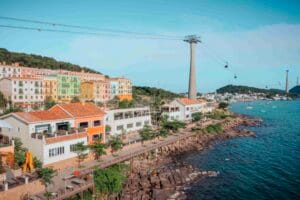 Dãy nhà nhiều màu kiểu Địa Trung Hải trên sườn đồi, trụ cáp treo cao giữa ảnh, lối đi gỗ chạy dọc bờ đá ra biển xanh