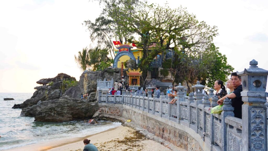 Miếu Dinh Cậu mái đỏ vàng trên ghềnh đá nhô ra biển, lan can đá chạm hoa văn, khách đứng ngắm biển