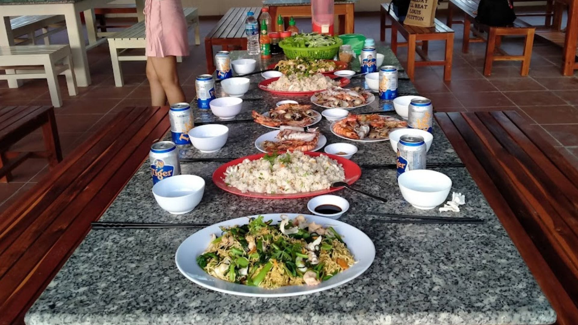 Bàn dài bày cơm, hải sản, rau và nước uống trong một quán gỗ