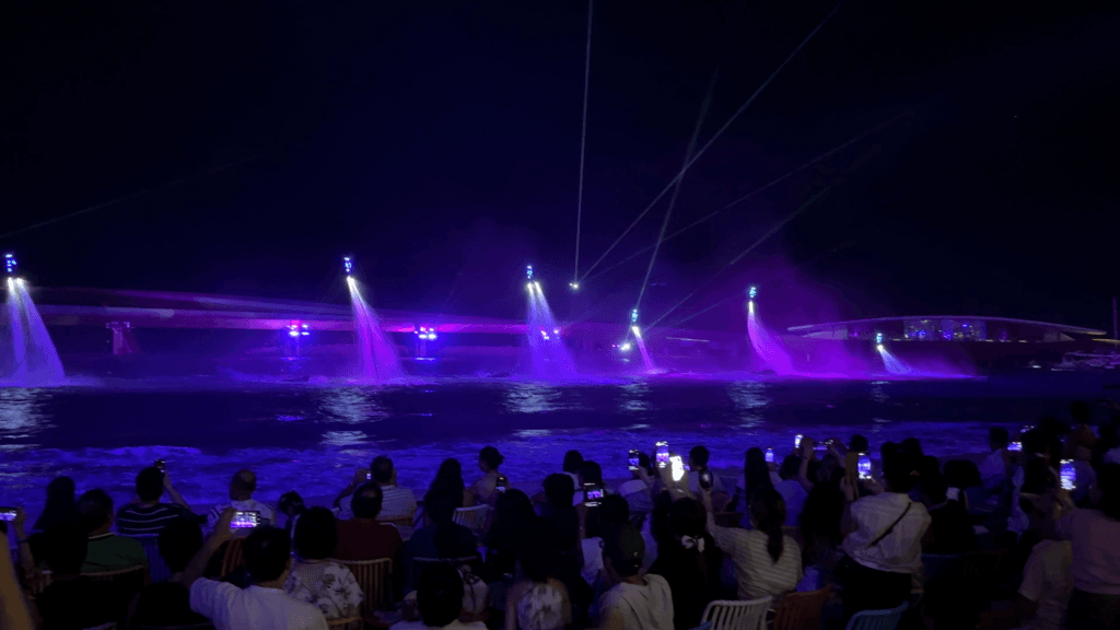 Khán giả ngồi kín khán đài sát bãi biển giơ điện thoại quay các cột nước và tia sáng tím trên mặt vịnh, phía sau là cây cầu lên đèn