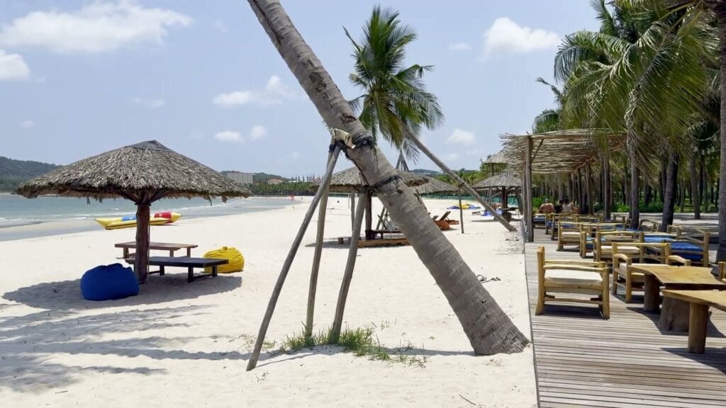 Bãi cát trắng có dù lá, ghế gỗ, bàn thấp và gối lười xanh vàng, hàng dừa nghiêng và sàn gỗ chạy dọc bãi, biển xanh bên trái